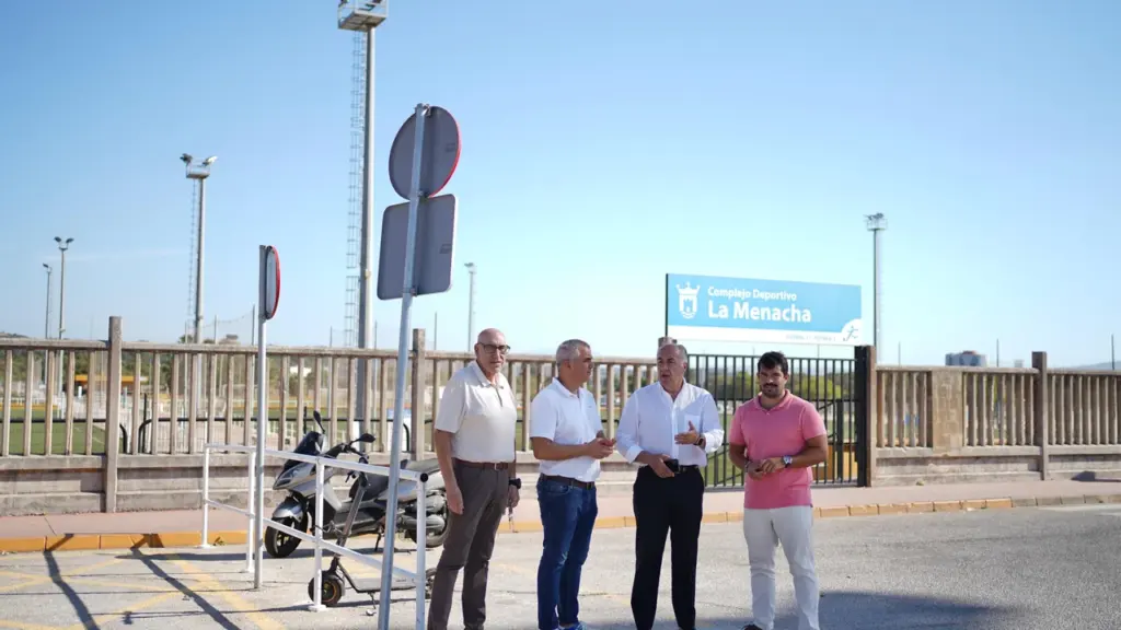 Cuatro hombres conversan de pie frente al cartel del Complejo Deportivo La Menacha en exterior.