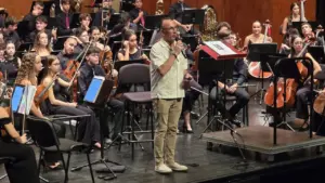 Un hombre con camisa clara habla ante un micrófono frente a una orquesta sinfónica completa.