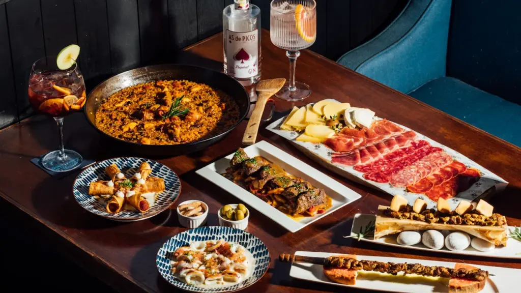 Mesa variada con platos de tapas, embutidos y bebida en restaurante español.