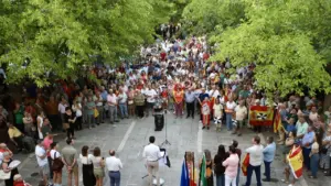 Gran multitud reunida en una plaza arbolada escuchando a un orador en un podio.
