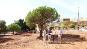 Cuatro hombres revisan planos en una obra de construcción con maquinaria pesada y árboles.